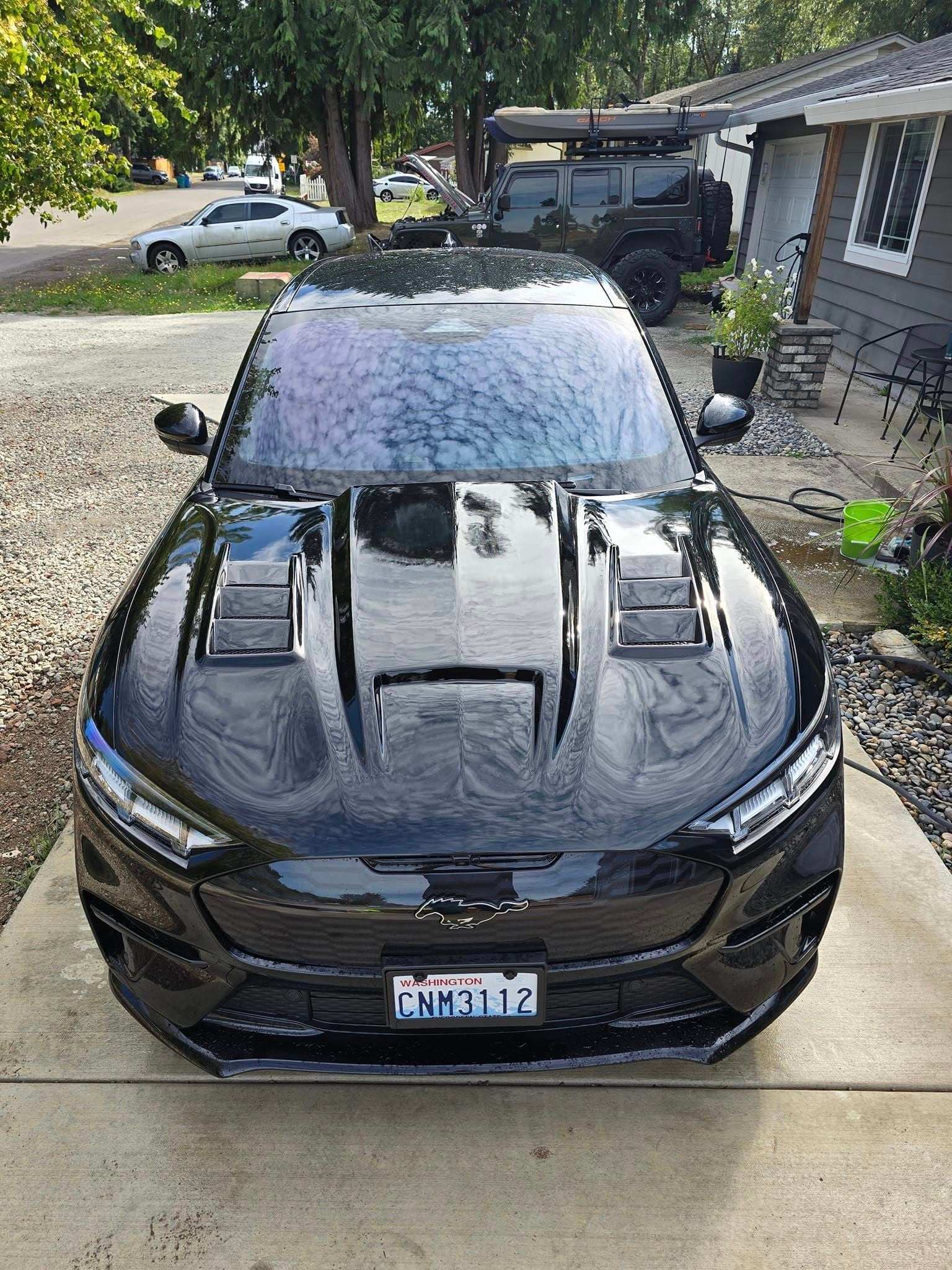 Front view of black Ford Mustang Mach-E with GT550 style wet carbon fiber hood featuring vents, parked in driveway