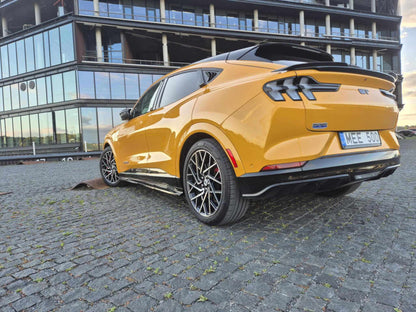Yellow Mustang Mach-E coupe with Spoiler V3 fender to fender installed on trunk, parked on cobblestone near modern glass building